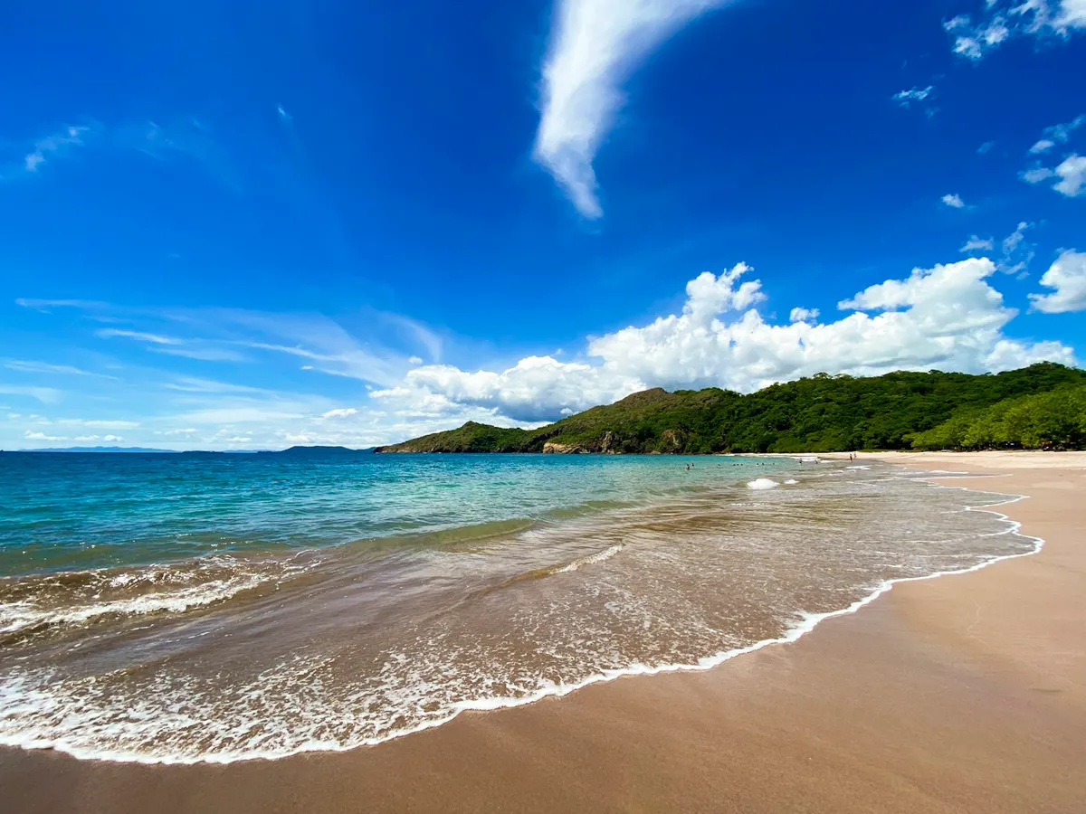 Calm tan-sand Guanacaste bay with a green headland under a blue sky near Playas del Coco, Costa Rica